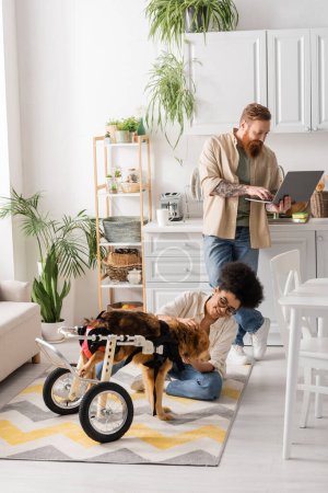 Smiling african american woman sitting near disabled dog and boyfriend using laptop in kitchen 