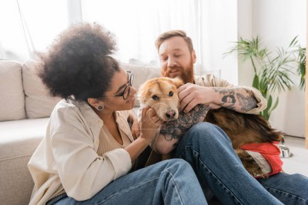 Cheerful multiethnic couple petting handicapped dog in living room 