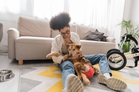 Young african american woman hugging handicapped dog near wheelchair and bowl on floor at home 