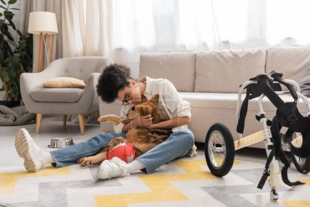 Young african american woman hugging disabled dog near wheelchair in living room 