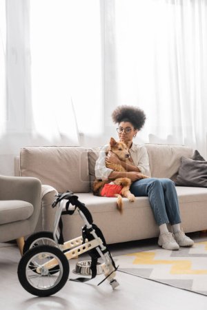 Joven mujer afroamericana abrazando perro discapacitado y mirando a la cámara en casa 