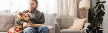 Bearded man petting handicapped dog while sitting on couch at home, banner 