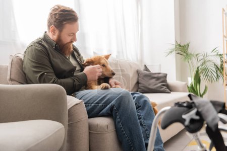 Bearded man sitting near disabled dog and blurred wheelchair in living room 