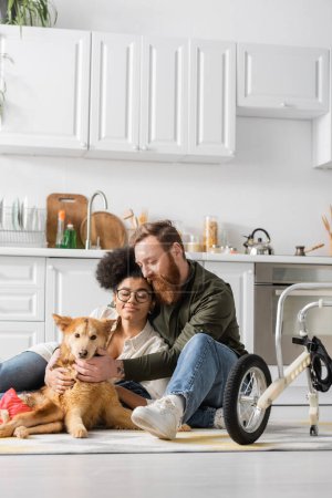 Multiethnic couple sitting near disabled dog on carpet near wheelchair in kitchen 