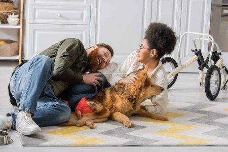 Happy bearded man spending time with african american girlfriend and disabled dog at home 