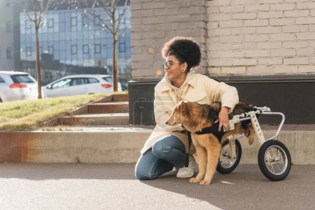 Smiling african american woman hugging dog with special needs on urban street in springtime 