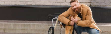 Bearded man hugging disabled dog in wheelchair on urban street, banner 