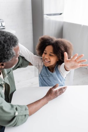 happy african american kid with curly hair smiling while stretching hands towards grandfather at home 