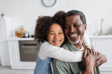 Foto de Retrato de chica afroamericana feliz con el pelo rizado sonriendo y abrazando abuelo alegre en casa - Imagen libre de derechos