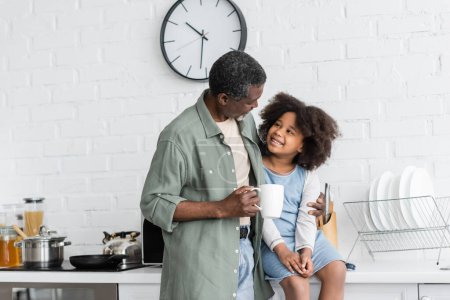 bärtiger afrikanisch-amerikanischer Großvater hält Tasse Kaffee in der Hand und umarmt glückliches Kind in der Küche 