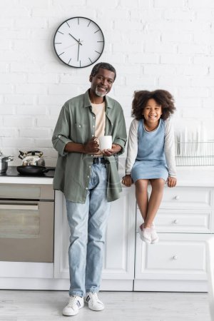 Afrikanisch-amerikanischer Großvater mittleren Alters hält Tasse mit Kaffee in der Nähe fröhliches Kind sitzt auf Küchenarbeitsplatte 
