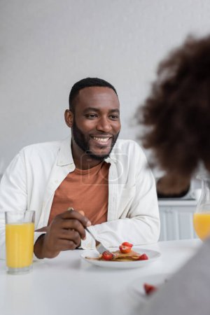 happy african american man having breakfast and looking at curly daughter in kitchen 