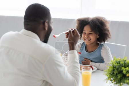happy african american girl having breakfast and looking at father eating strawberry on blurred foreground 