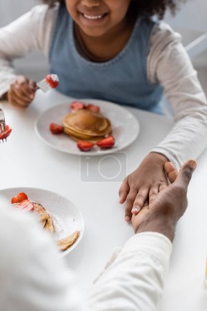 abgeschnittene Ansicht eines glücklichen afrikanisch-amerikanischen Mädchens, das beim Frühstück Händchen hält mit seinem Vater 