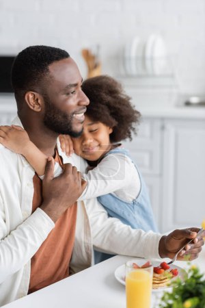 curly african american girl smiling while hugging happy father during breakfast 
