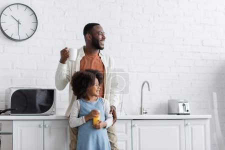 happy african american man holding cup of coffee and hugging cheerful daughter with glass of orange juice 
