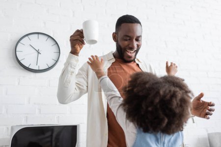 curly african american kid stretching hands towards happy father with cup of coffee 