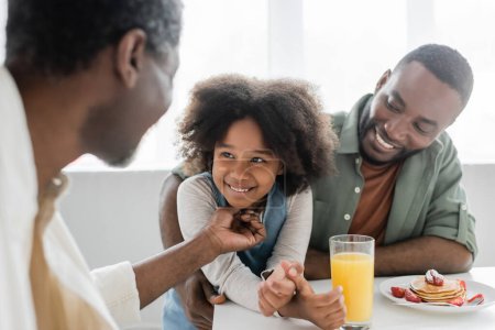 Afrikanisch-amerikanischer Großvater kitzelt glückliche Enkelin neben fröhlichem Sohn beim Familienfrühstück 
