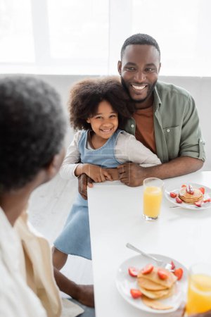 Téléchargez les photos : Heureux homme afro-américain étreignant fille bouclée et regardant père pendant le petit déjeuner familial - en image libre de droit
