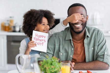 lockiges afrikanisch-amerikanisches Mädchen mit Glückwunschkarte mit dem Schriftzug "Happy Vatertag", während sie die Augen von Papa bedeckt 