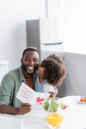 curly african american kid kissing cheek of happy father with greeting card on fathers day 