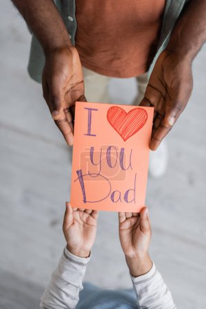 von oben Ansicht beschnitten afrikanisch-amerikanisches Kind hält Grußkarte mit Ich liebe dich Papa Schriftzug in der Nähe Mann am Vatertag 