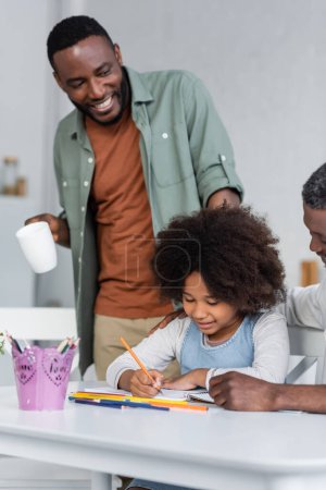 happy african american man holding cup and standing near daughter drawing on paper near grandpa 