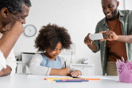 hombre afroamericano feliz tomando fotos en el teléfono inteligente mientras su hija y su padre dibujan juntos 