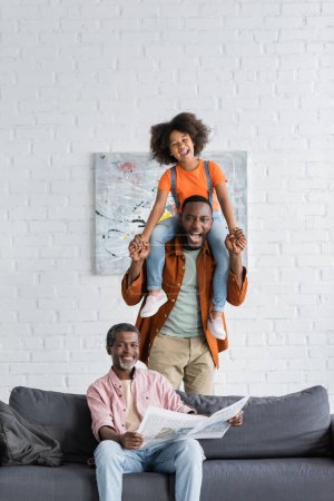 Overjoyed african american man and kid having fun near grandparent with newspaper on couch at home 
