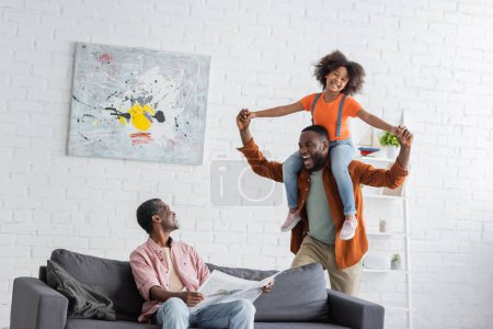 African american grandparent holding newspaper near son and granddaughter having fun at home 