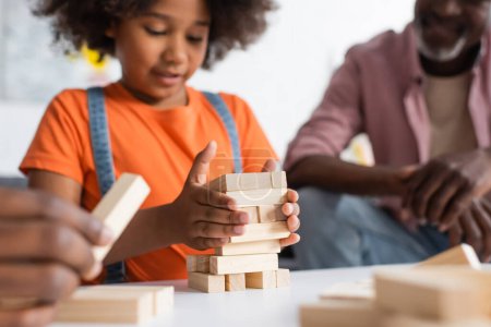 Téléchargez les photos : Enfant afro-américain flou jouant à un jeu de blocs de bois près des parents à la maison - en image libre de droit