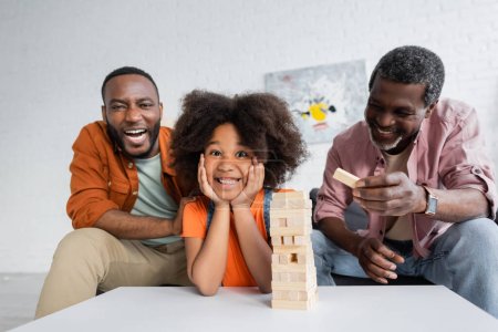 Fröhliches afrikanisch-amerikanisches Kind blickt beim Holzklötzchenspiel zu Hause in die Kamera neben Opa und Vater 