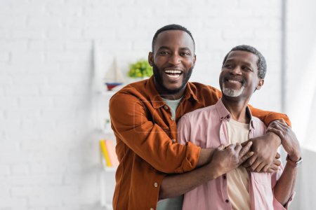 Téléchargez les photos : Joyeux homme afro-américain embrassant papa mature et regardant la caméra à la maison - en image libre de droit