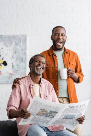 Cheerful african american man holding cup of coffee near dad with newspaper at home 