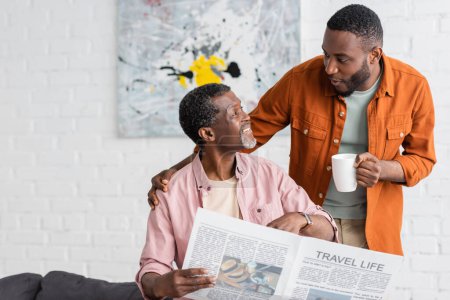 African american man holding cup of coffee and talking to father with newspaper at home 
