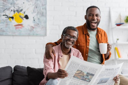 Despreocupado hombre afroamericano con taza abrazando padre maduro con periódico en la sala de estar 
