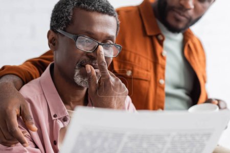 Mature african american man in eyeglasses reading newspaper near blurred son at home 