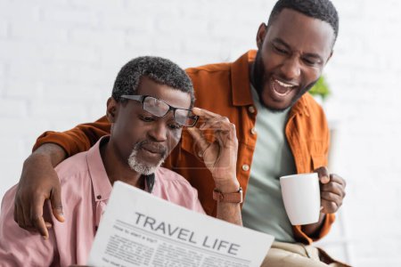 Excited african american man holding coffee cup while father reading newspaper at home 