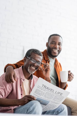 Fröhlicher afrikanisch-amerikanischer Mann hält eine Tasse Kaffee in der Hand und umarmt seinen reifen Vater mit Reisenachrichten zu Hause 