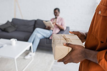 African american man holding gift box during father day celebration near blurred dad  at home