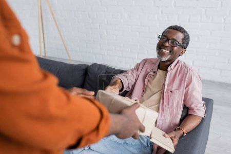 Téléchargez les photos : Joyeux homme afro-américain avec livre prenant boîte cadeau de fils pendant la fête des pères à la maison - en image libre de droit