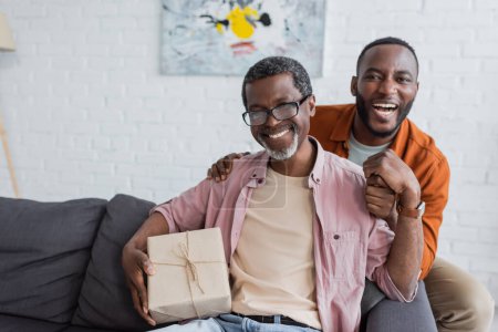 Fröhlicher afrikanisch-amerikanischer Mann mittleren Alters hält Geschenk in der Nähe seines Sohnes während der Vatertagsfeier zu Hause 