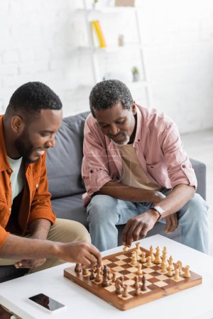 Hombre afroamericano jugando ajedrez con hijo sonriente cerca de teléfono inteligente en casa 