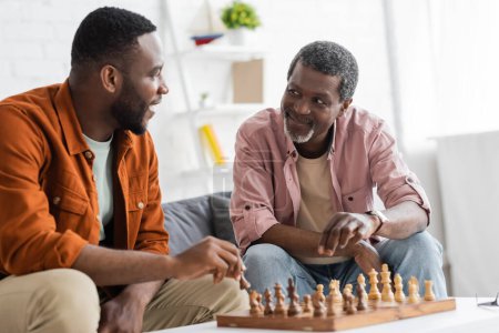 Sonriente hombre afroamericano jugando ajedrez con hijo adulto en la sala de estar 