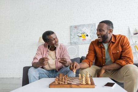 Alegre hombre afroamericano jugando ajedrez con padre maduro cerca de teléfono inteligente con pantalla en blanco en casa 