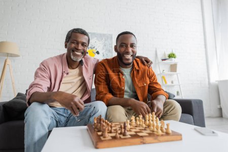 Sonriente hombre afroamericano con anteojos abrazando hijo y mirando a la cámara cerca de ajedrez y teléfono inteligente en casa 