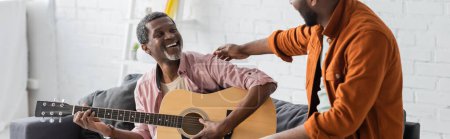 Alegre hombre afroamericano tocando la guitarra acústica cerca de hijo en casa, pancarta 