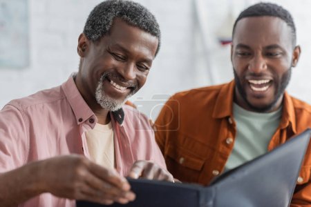 Carefree african american man pointing at blurred photo album near son at home 