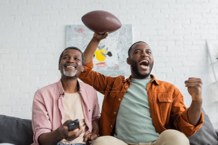Emocionado hombre afroamericano sosteniendo pelota de rugby cerca de papá con controlador remoto en la sala de estar 