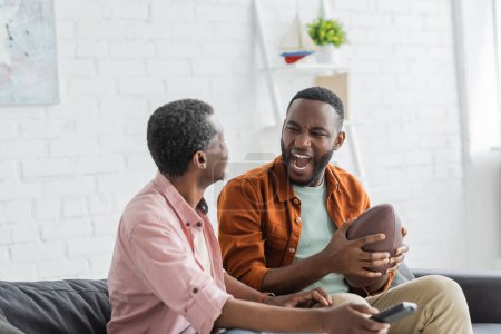 Excited african american man holding rugby ball near mature dad with remote controller at home 